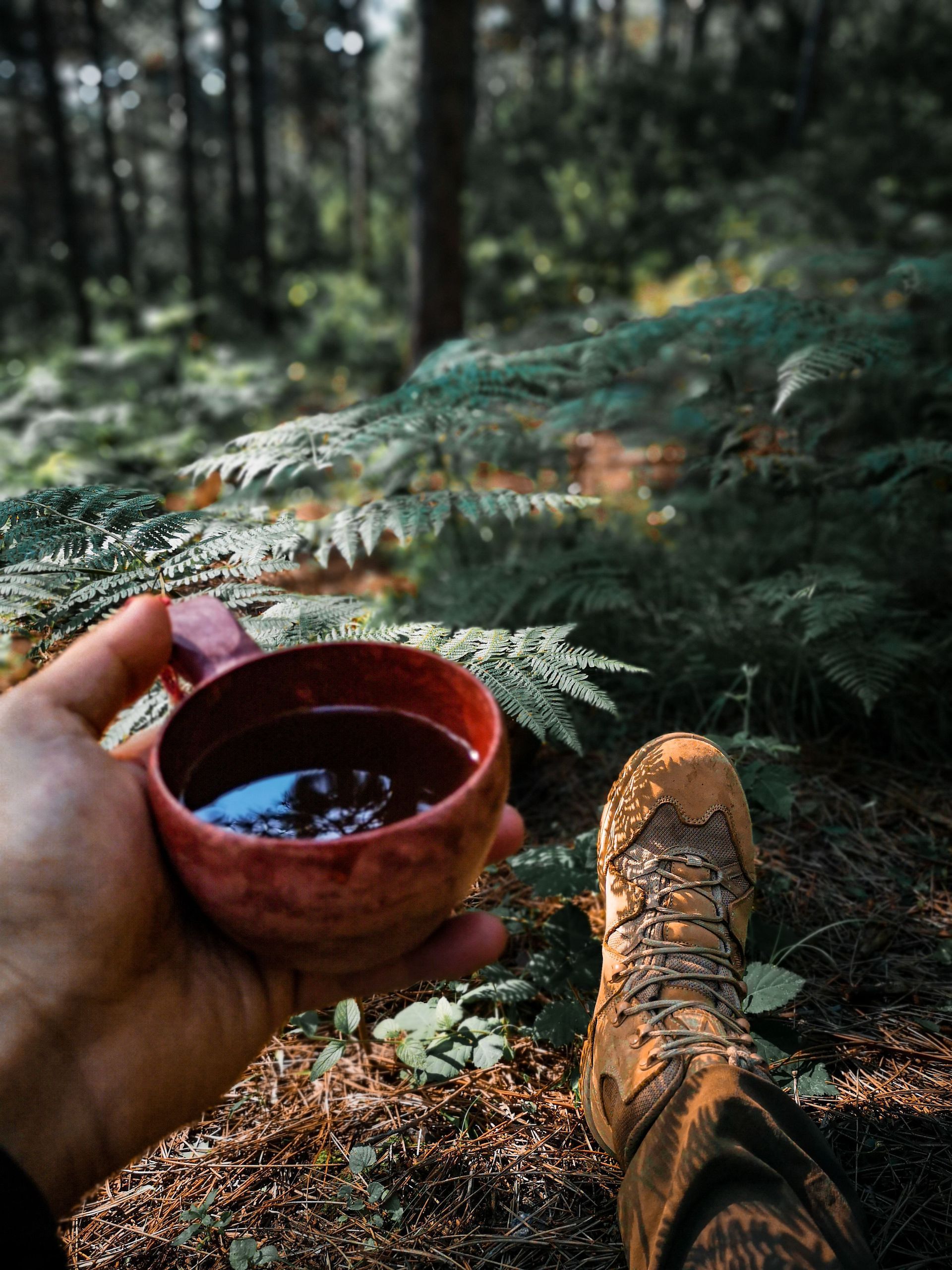 Hand holding a small red cup beside a hiking boot in a forest with moss and ferns