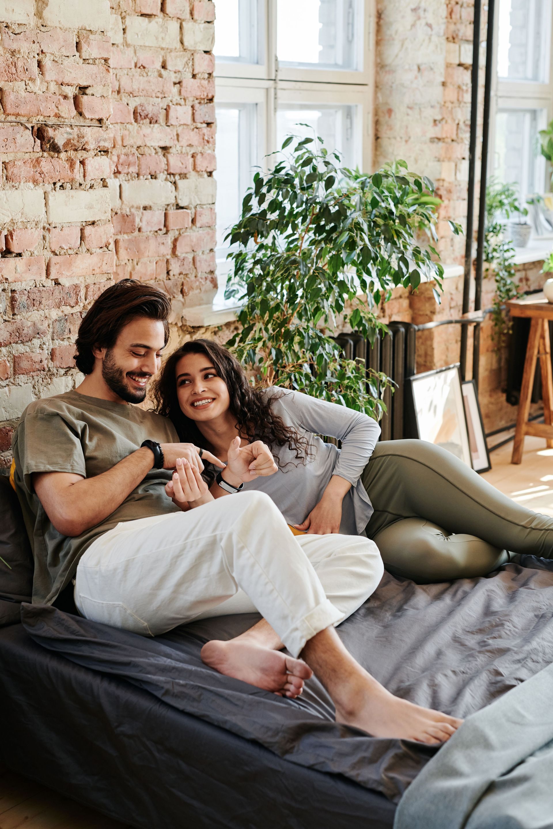 Couple relaxing on a bed in a bright loft, smiling and holding mugs near a brick wall and large windows.