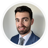 Man in blue suit, smiling, in front of a light gray wall.