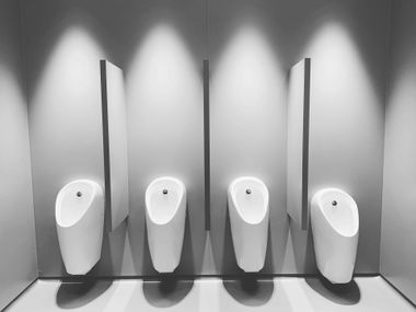 Four white urinals in a public restroom stall with gray walls and bright overhead lighting
