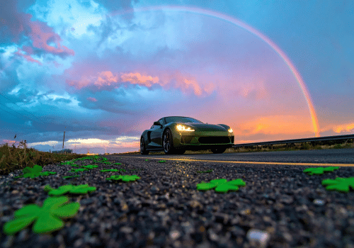 Sports car driving on a road at sunset with a rainbow overhead and shamrocks on the ground.