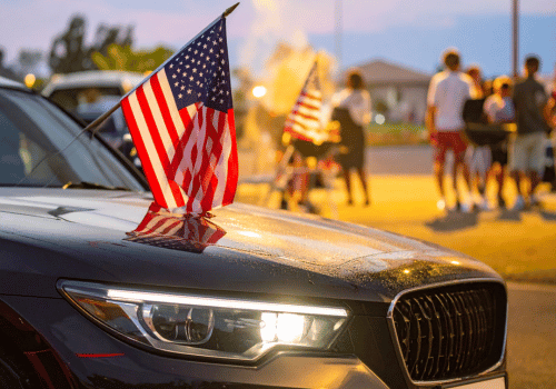 Close-up of a car with an American flag attached, parked near people gathered outdoors at dusk.