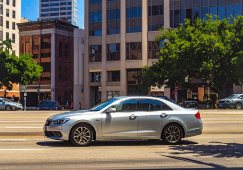 Silver sedan driving through a downtown city street with office buildings, trees, and bright midday
