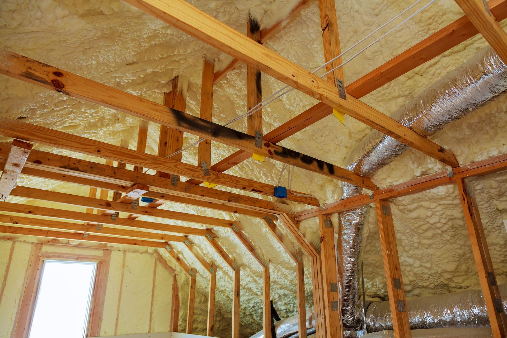 Attic interior with wooden rafters and spray foam insulation, window, and electrical boxes.