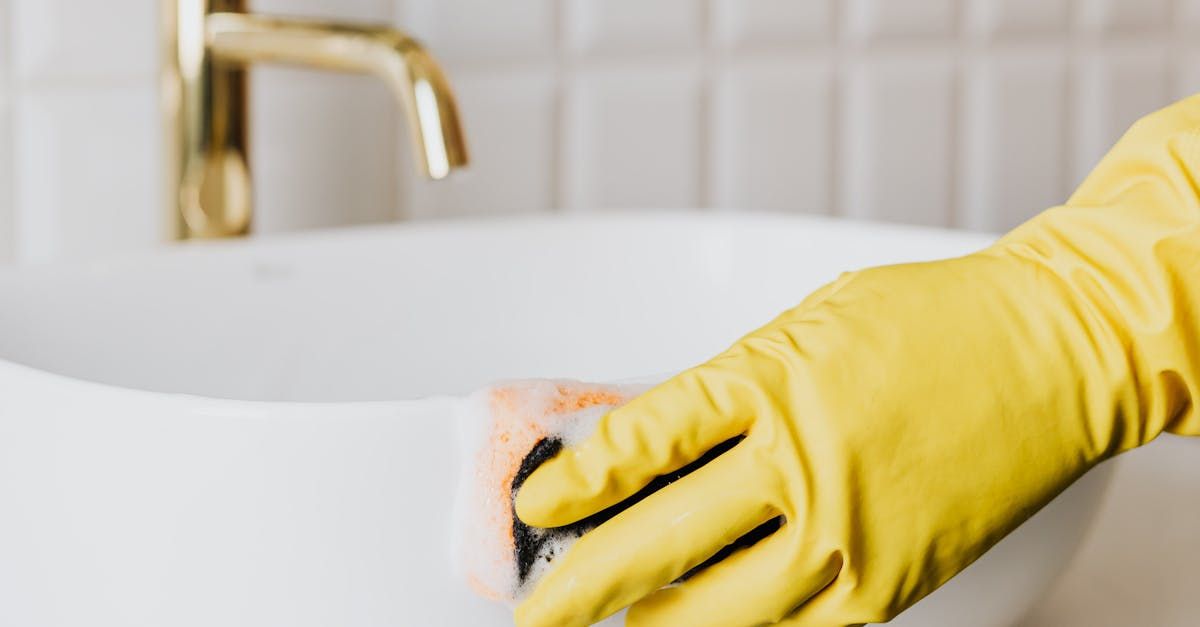 A person wearing yellow gloves is cleaning a bathroom sink with a sponge.