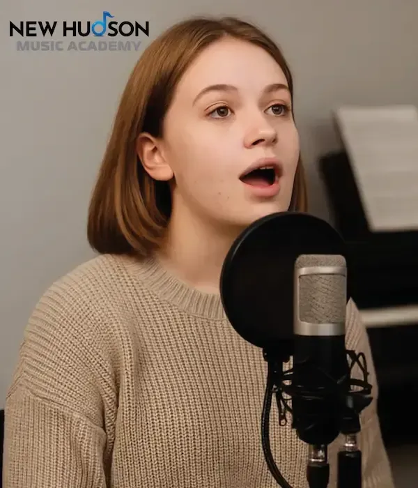A person singing into a microphone in a music studio, with the New Hudson Music Academy logo in the top left corner.