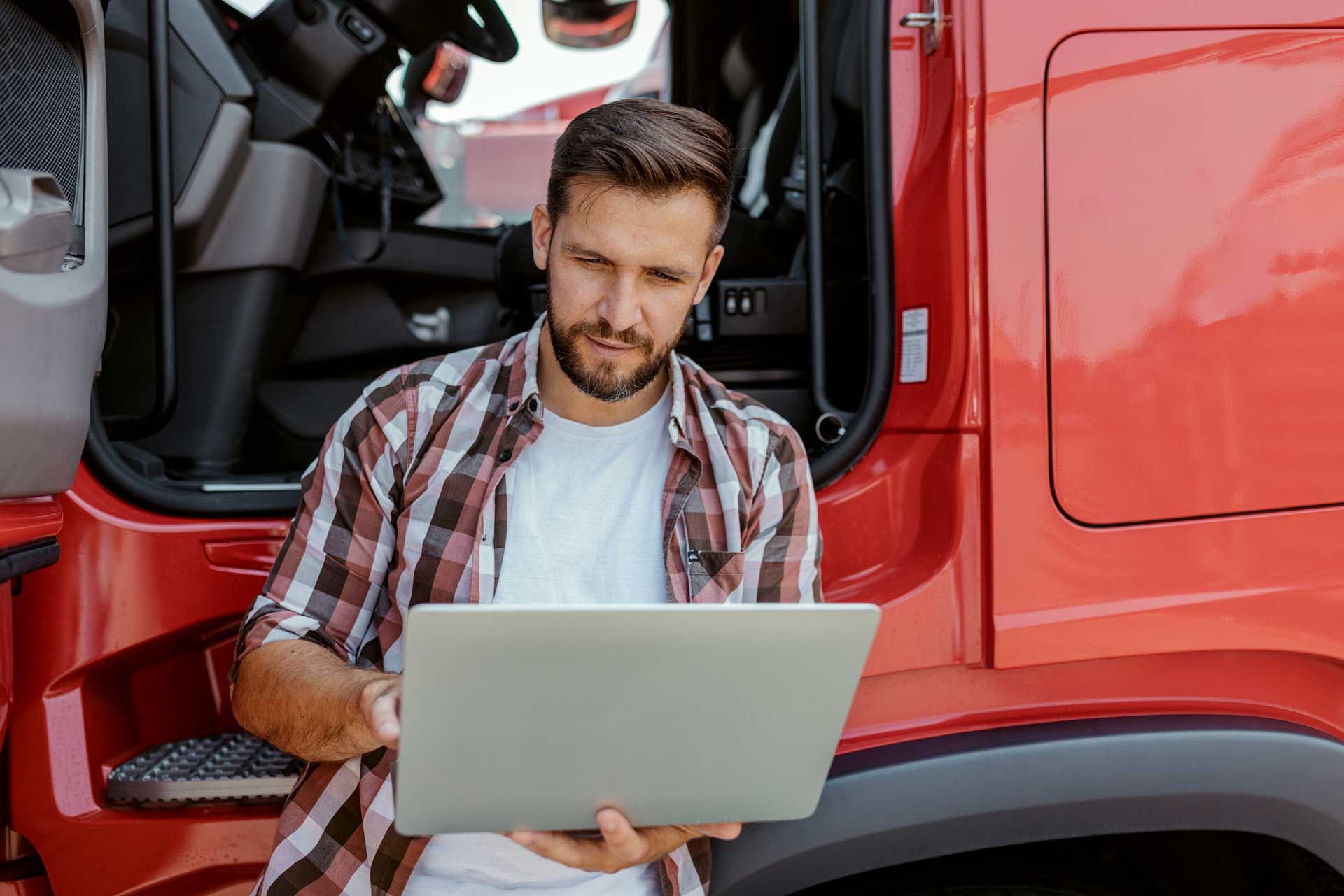 A man is sitting in front of a red truck using a laptop computer.