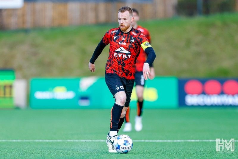 Knattspyrnumaður í rauðum og svörtum treyju dribblar boltann á vellinum í leik