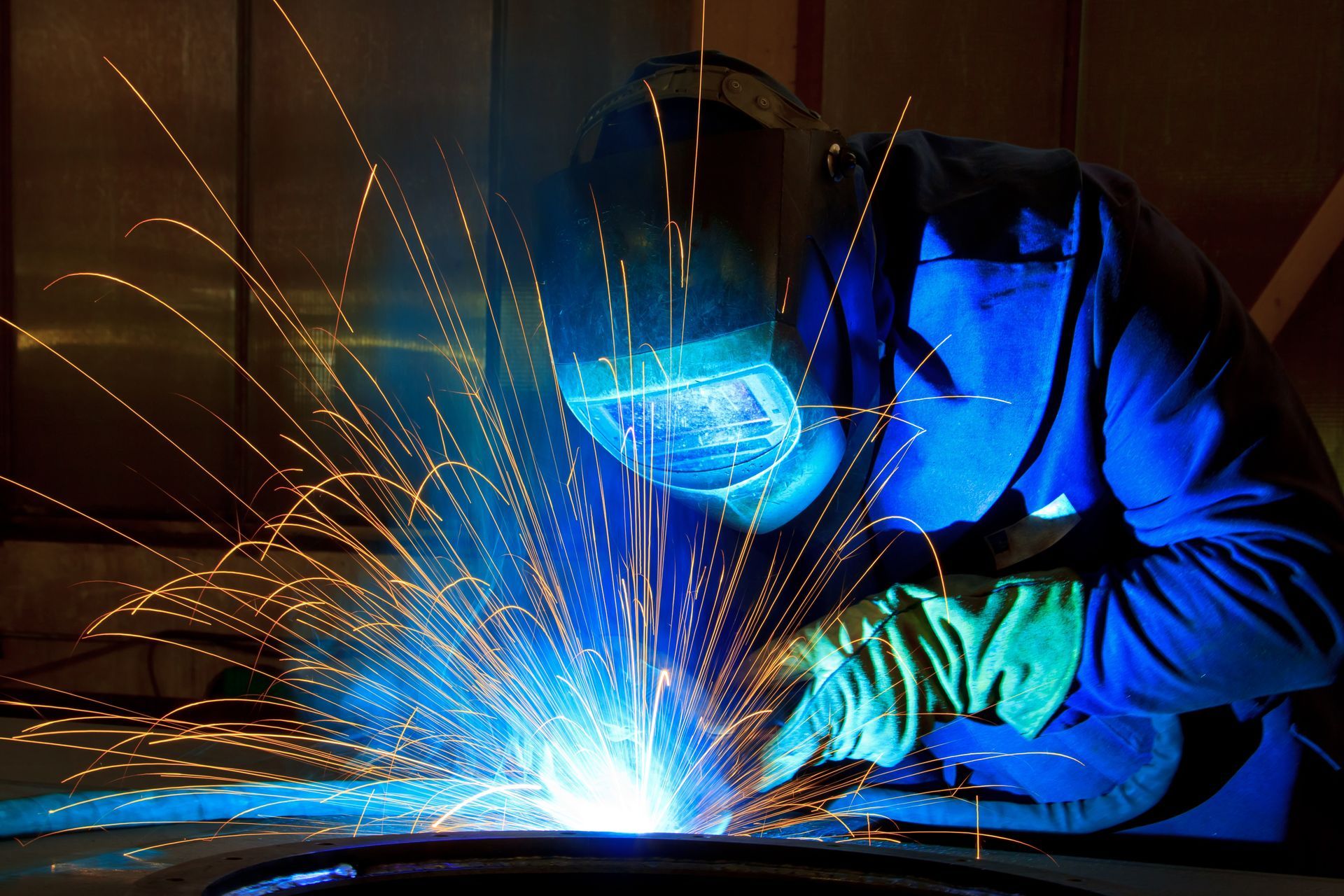 A welder wearing protective gear and a welding helmet, actively welding metal.