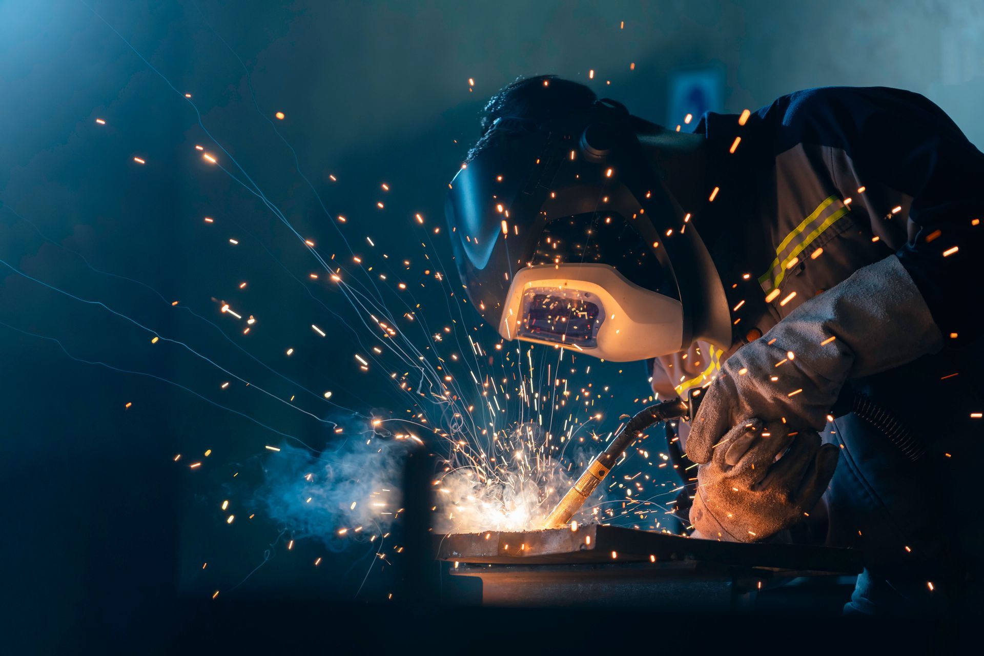 A welder wearing a dark helmet and gloves welds steel in a shadowy industrial setting.