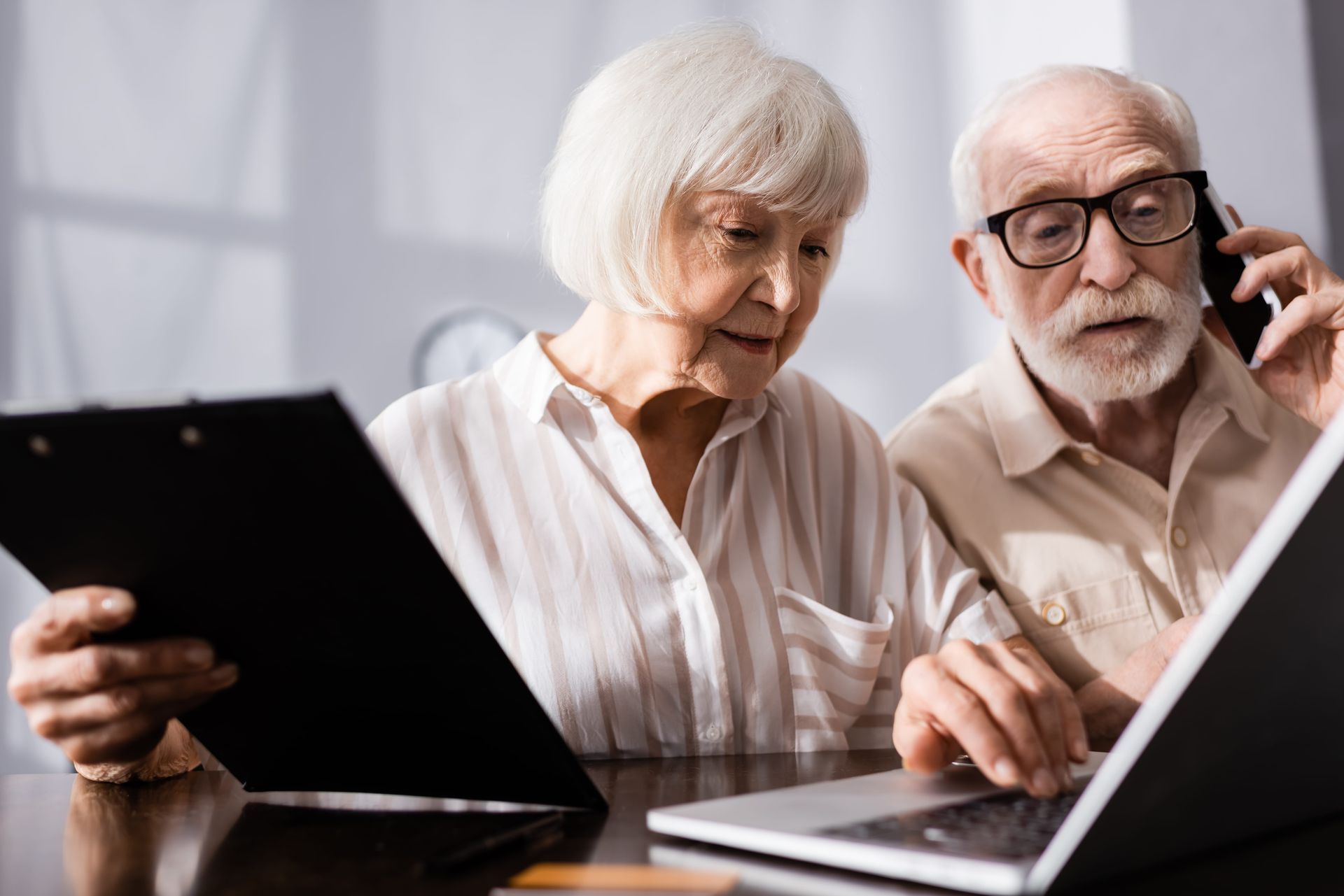 Two older adults reviewing a laptop together, one holding a tablet, in a bright indoor setting