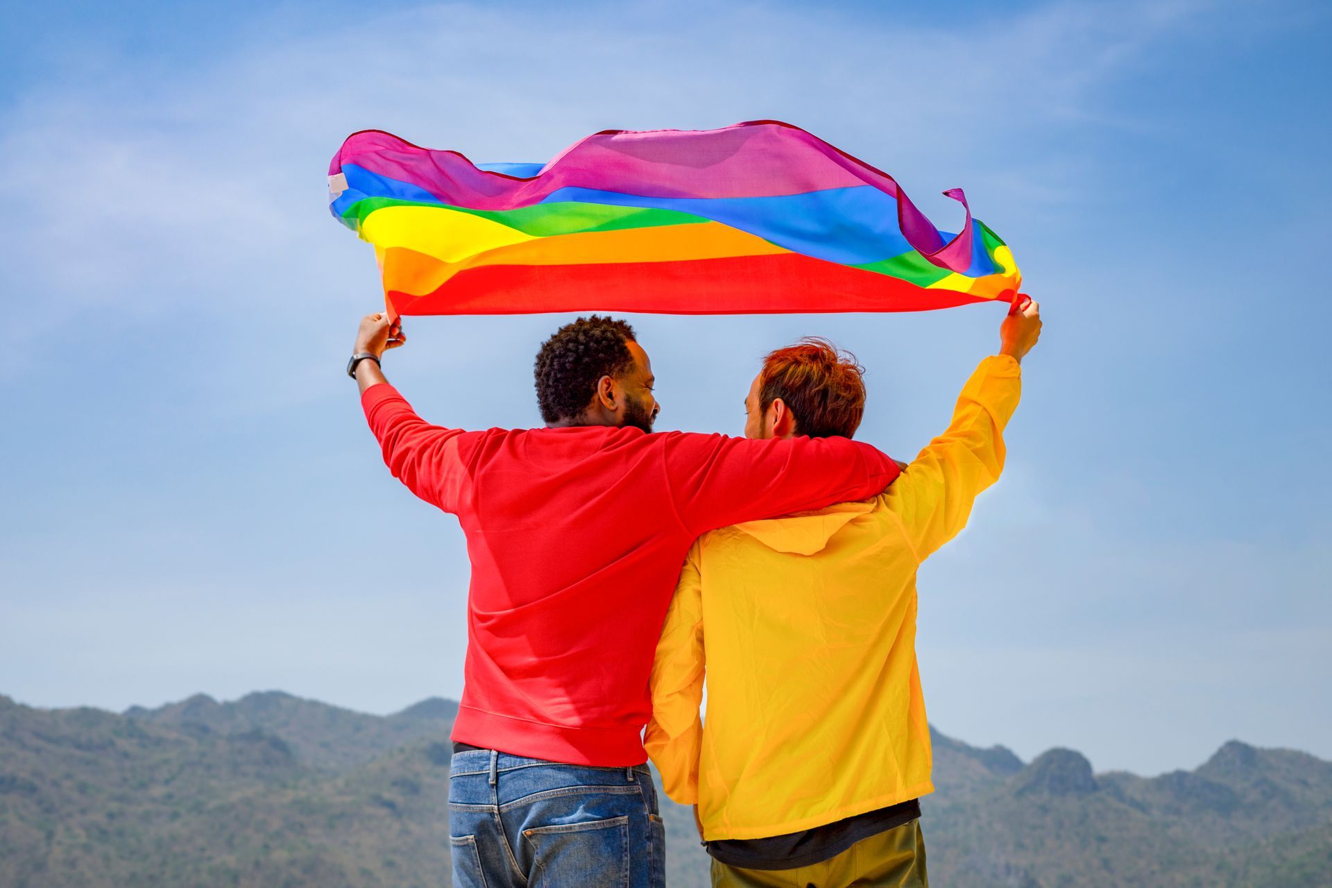 Two men embrace, holding a rainbow flag aloft against a clear sky and mountainous backdrop.