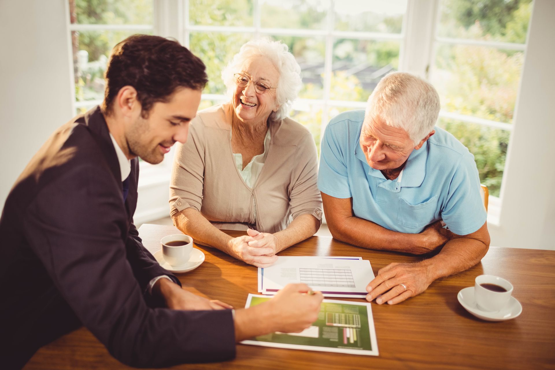 Real estate agent showing house plan to elderly couple at a table, smiling, sunlight.