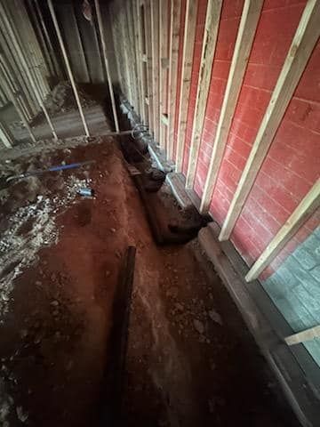 Trench dug in a room under construction, along red brick wall and wooden framing.