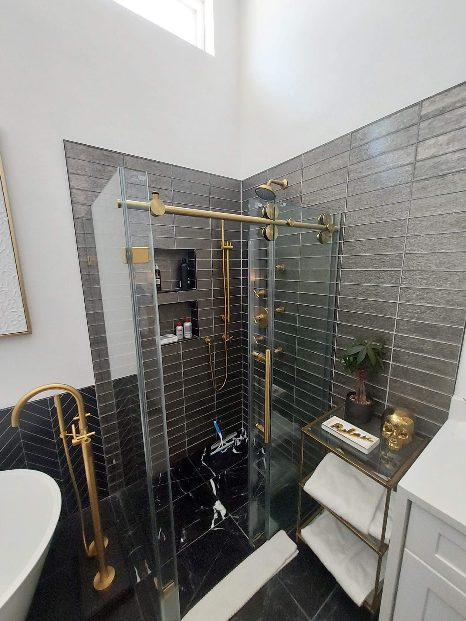 Luxurious bathroom with a glass shower, gold fixtures, black and silver tile, and a white tub.