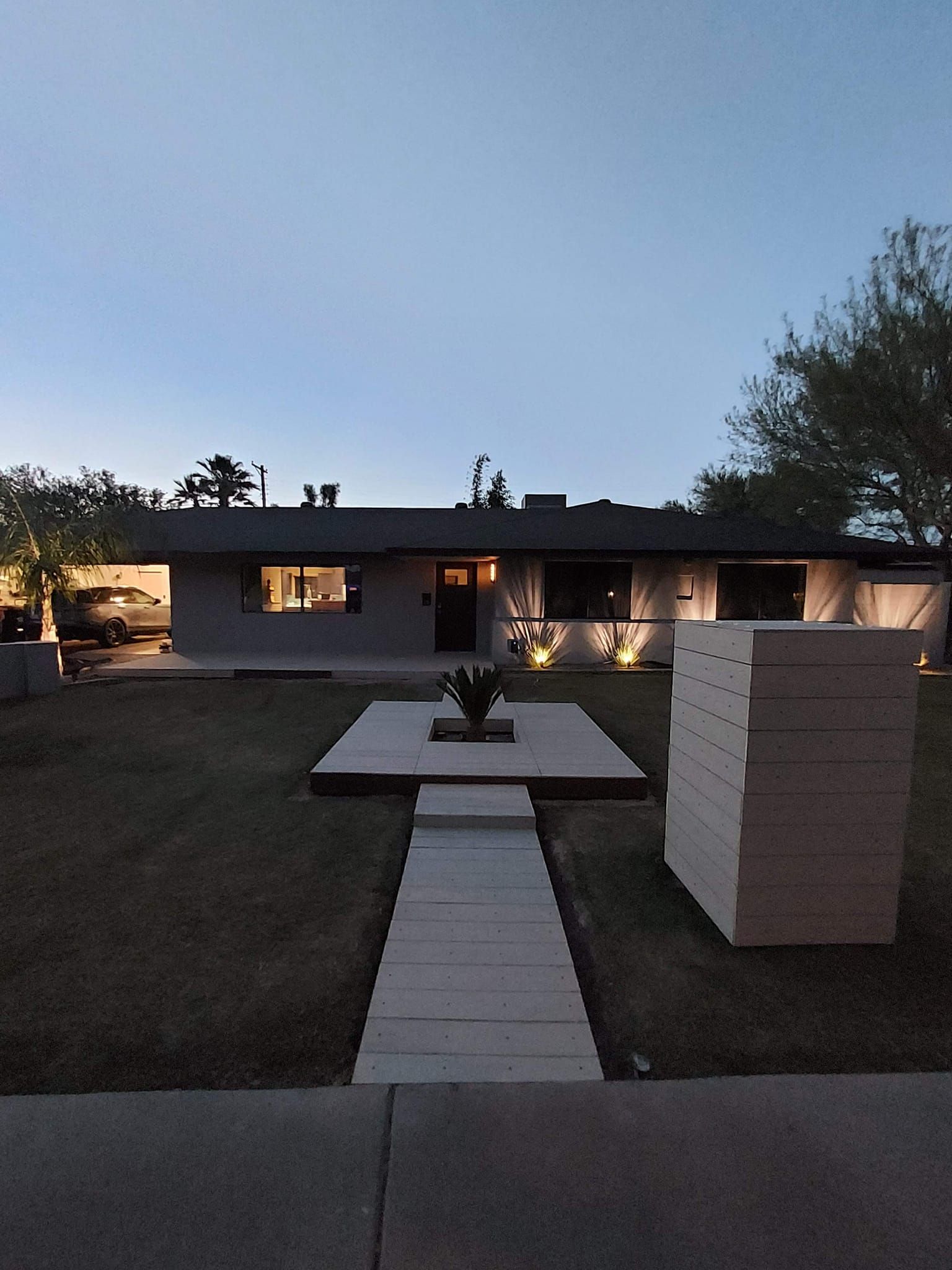 A house at dusk with a lighted pathway and stacked wood on the right.