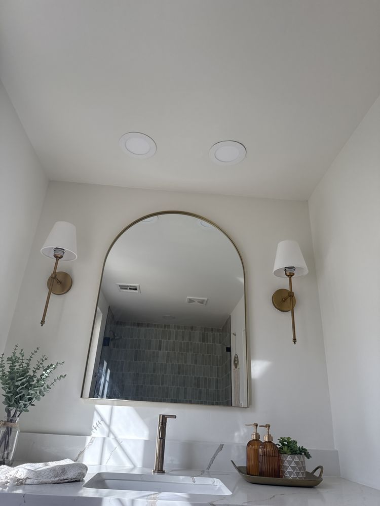 Bathroom with arched mirror, gold sconces, overhead lights, white walls, and countertop.