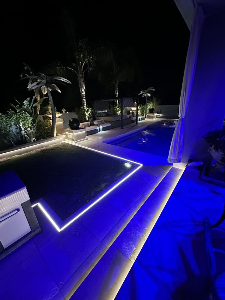 Nighttime view of a luxury backyard with a pool, illuminated by blue and white lights.