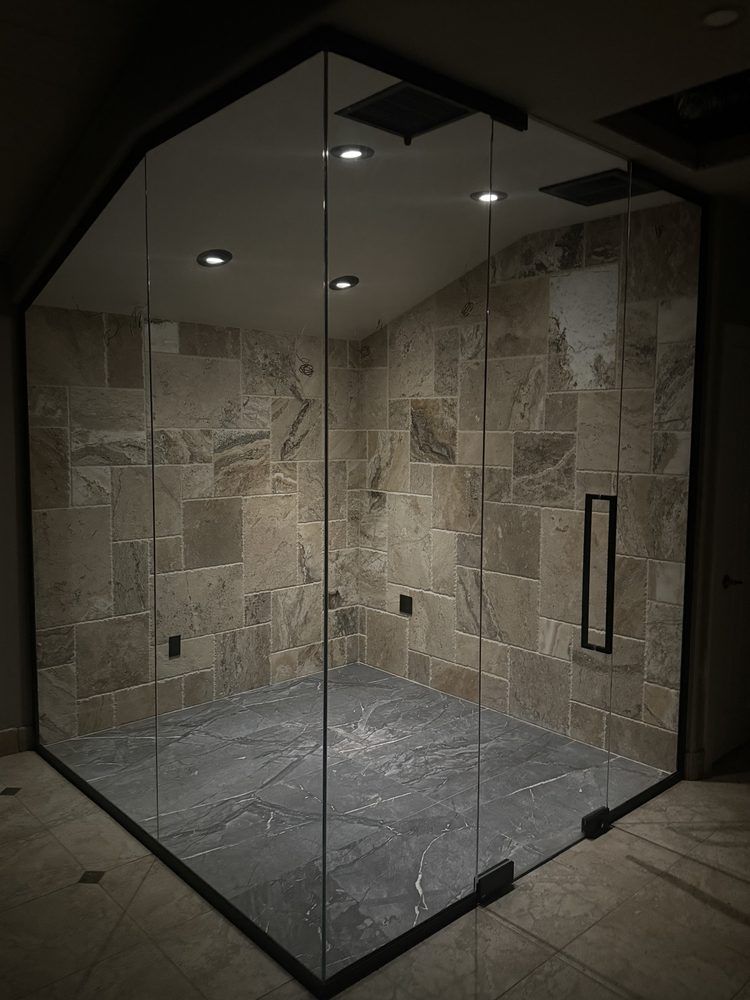 Glass-enclosed shower with black framing, beige stone tile walls, and gray stone floor tiles.