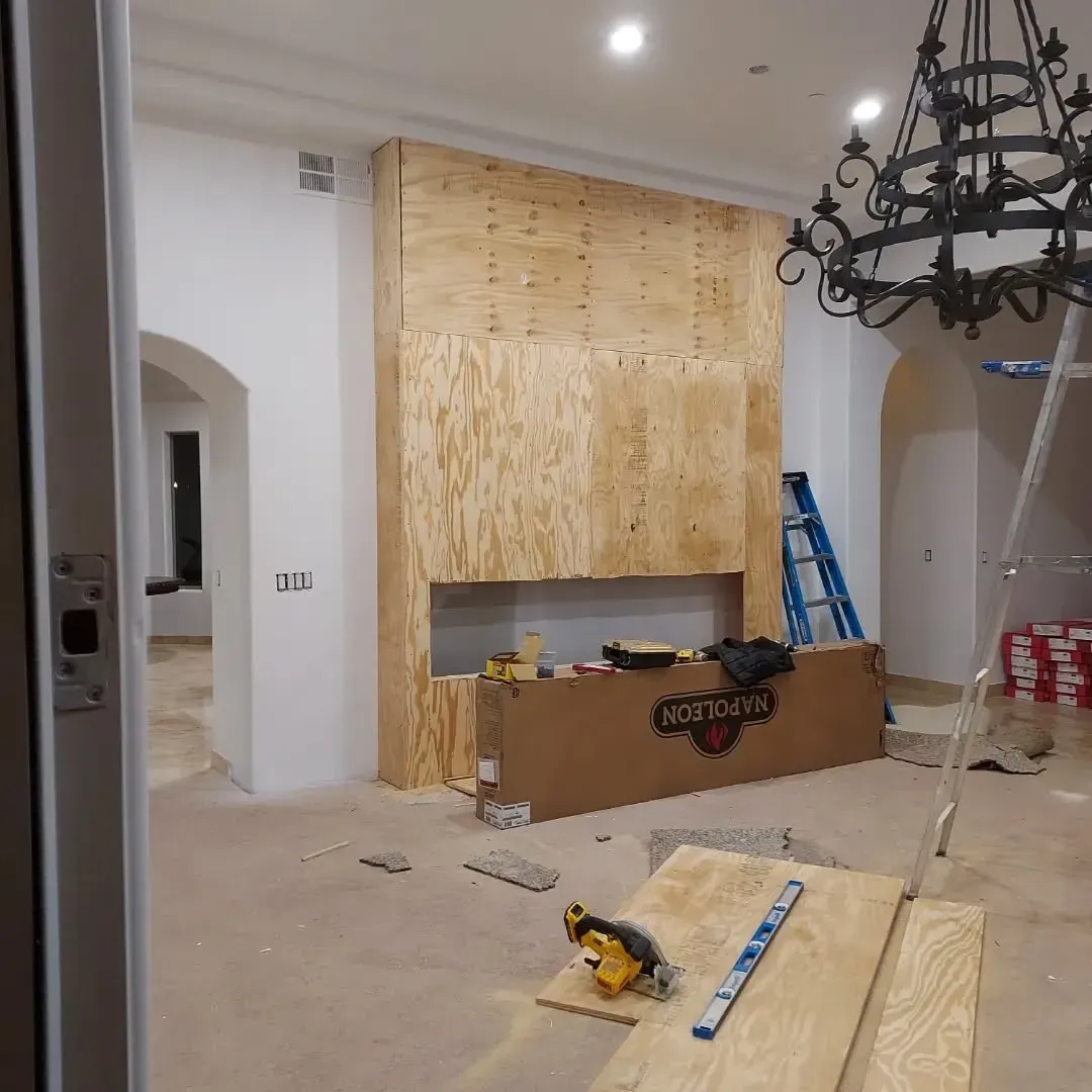 Room with unfinished fireplace, covered in plywood. Tools and debris scattered.