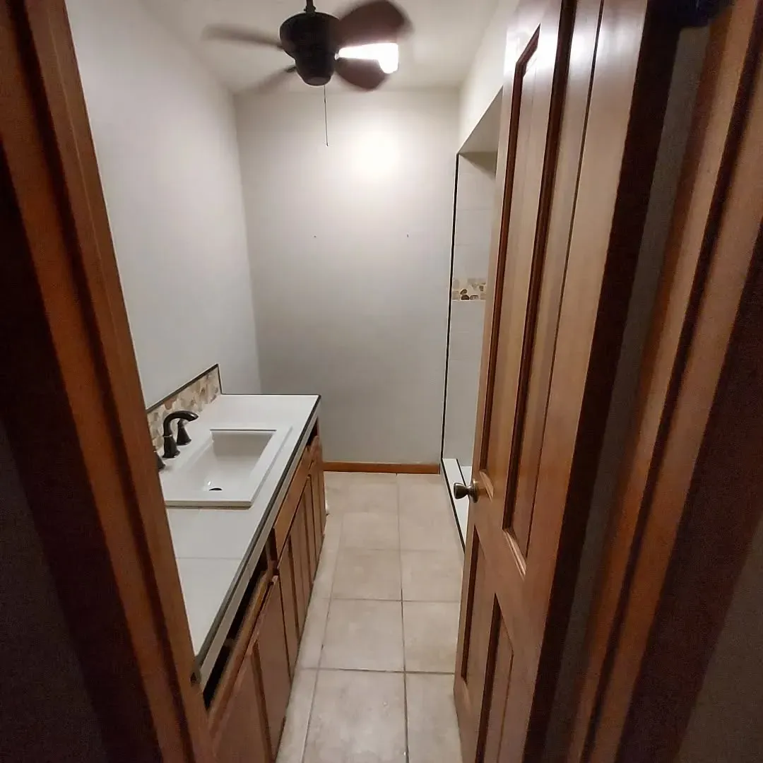 Narrow bathroom with sink, wooden cabinets, door, and ceiling fan.