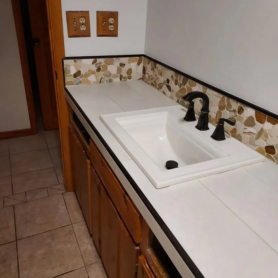 Bathroom sink with white countertop, brown cabinets, and stone backsplash.