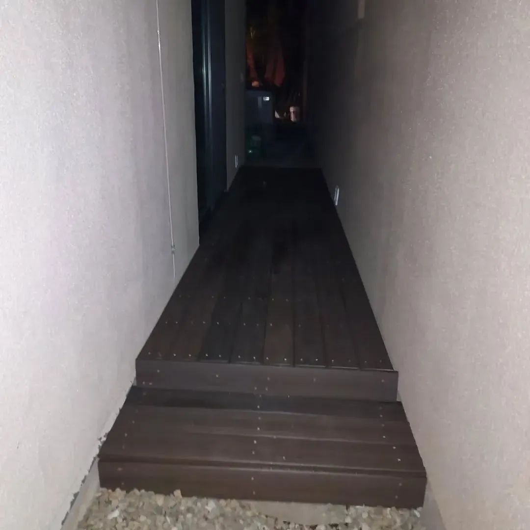 Narrow, dark pathway between white walls, wooden steps lead to a dark area with a raised walkway.