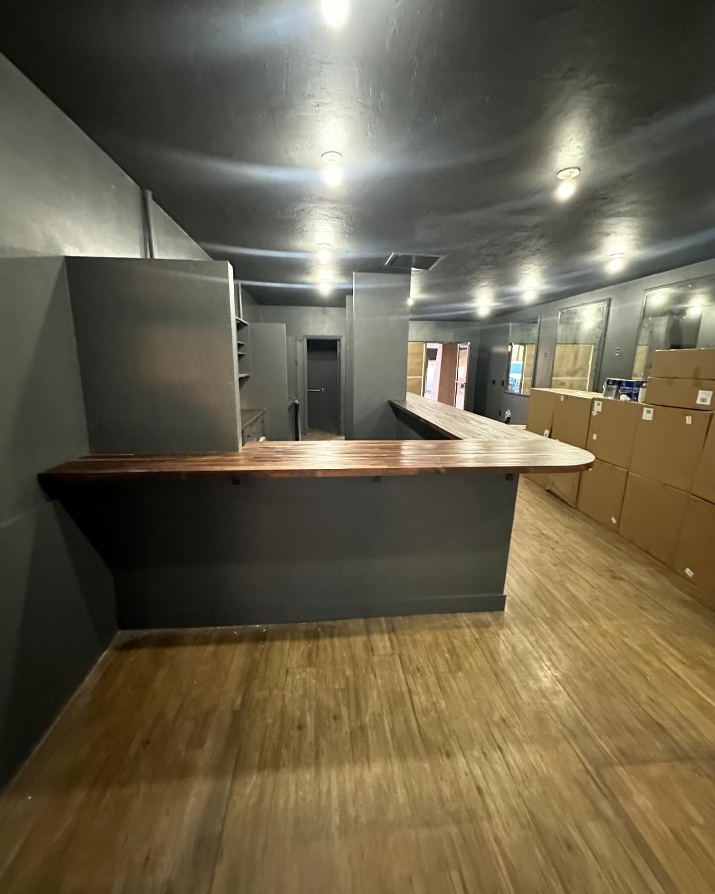 Empty bar interior with wooden counter, dark gray walls, and wooden floor. Boxes line the right side.