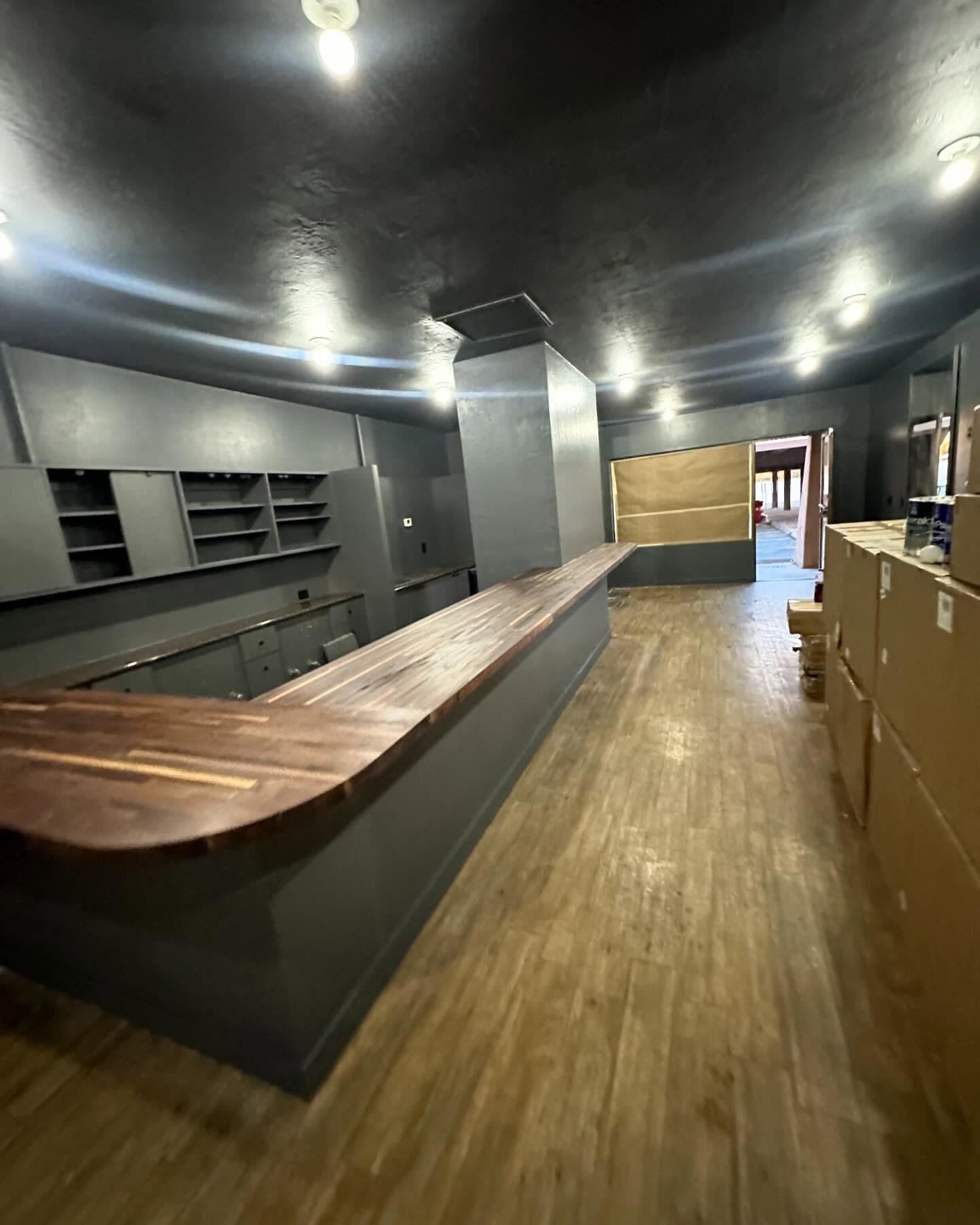 Empty bar with wooden counter, shelves, and gray walls. Cardboard boxes line the right.
