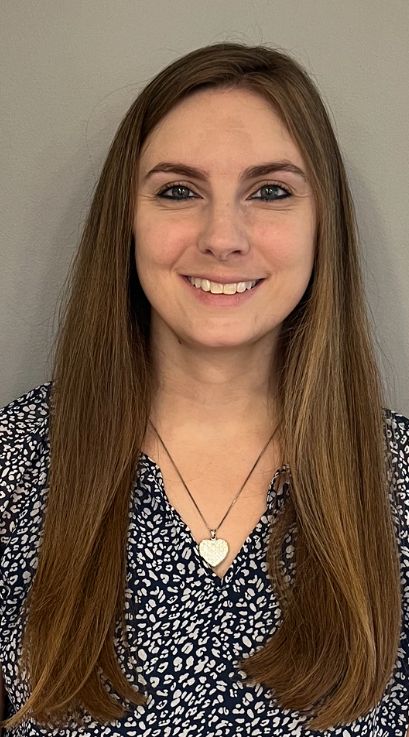 A woman in a red shirt and necklace is standing in front of a tree.