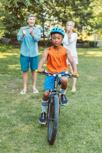 A young boy is riding a bike in a park while his parents applaud.