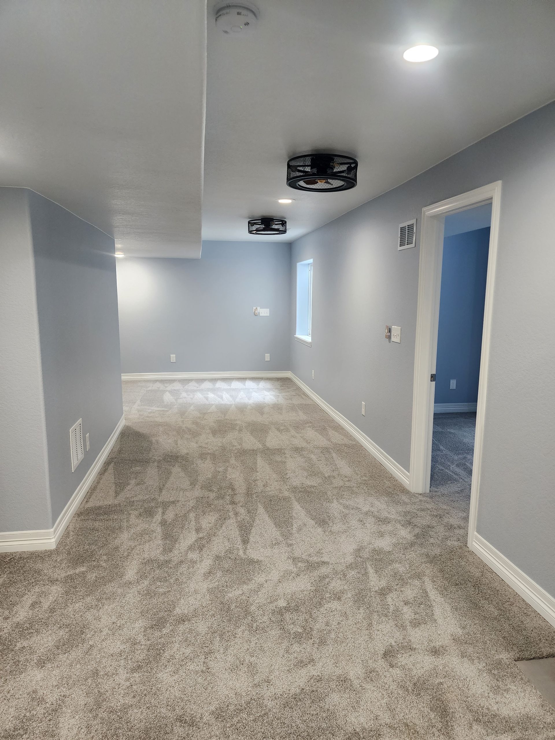A newly finished basement room with gray carpet, walls, and two dark ceiling fans. A doorway leads to another room.