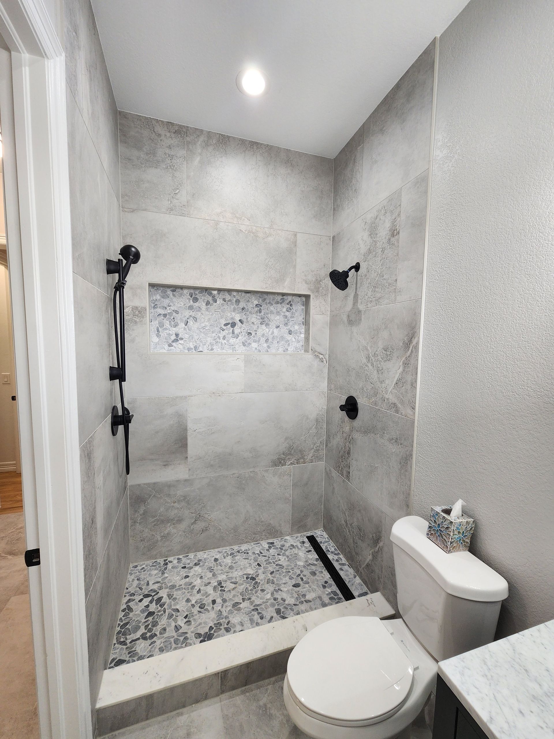 A modern bathroom with a gray tiled shower, black fixtures, and a toilet with a tissue box.
