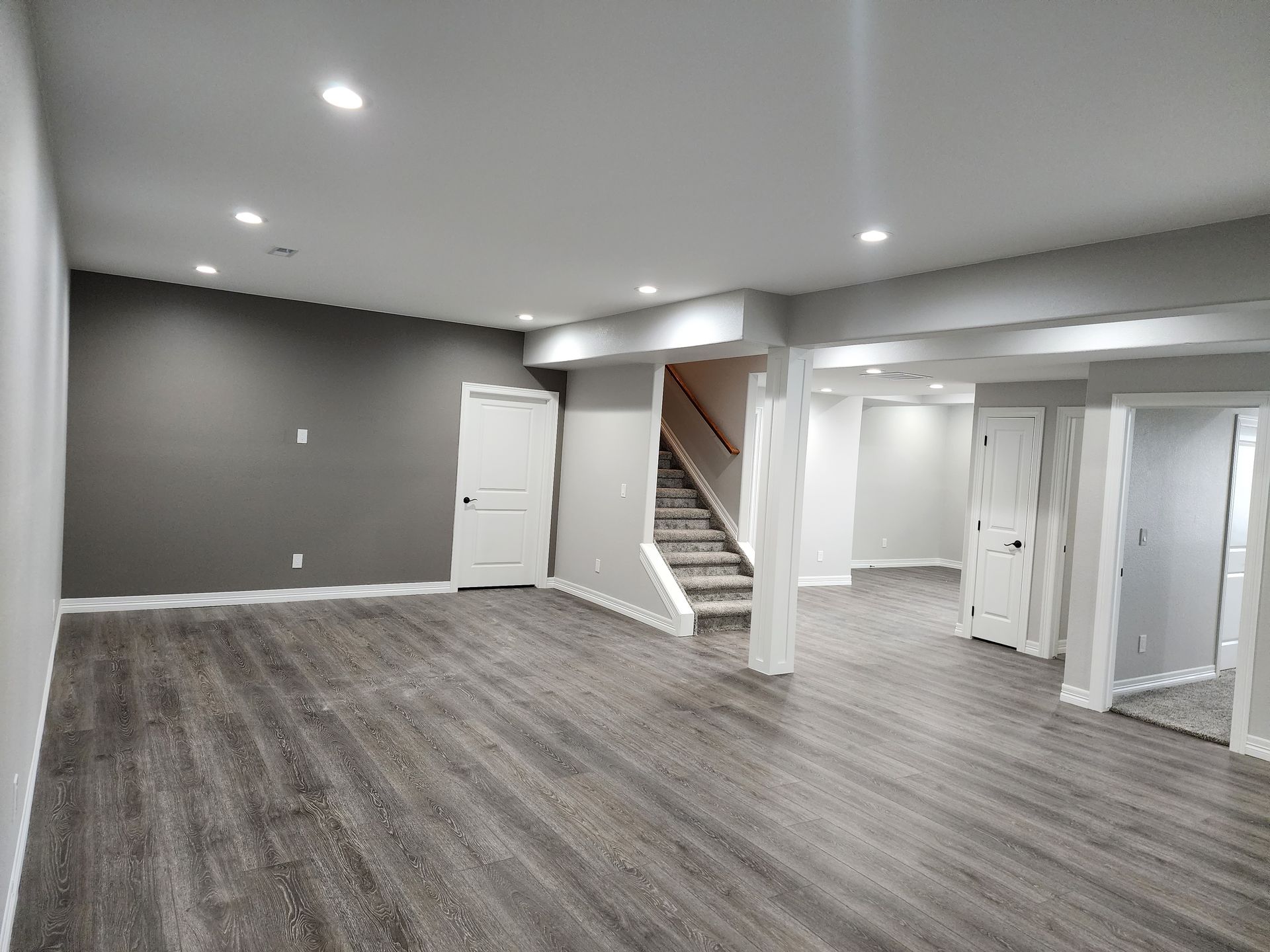 Empty basement with gray walls, wood-look floor, recessed lighting, stairs, and white doors.