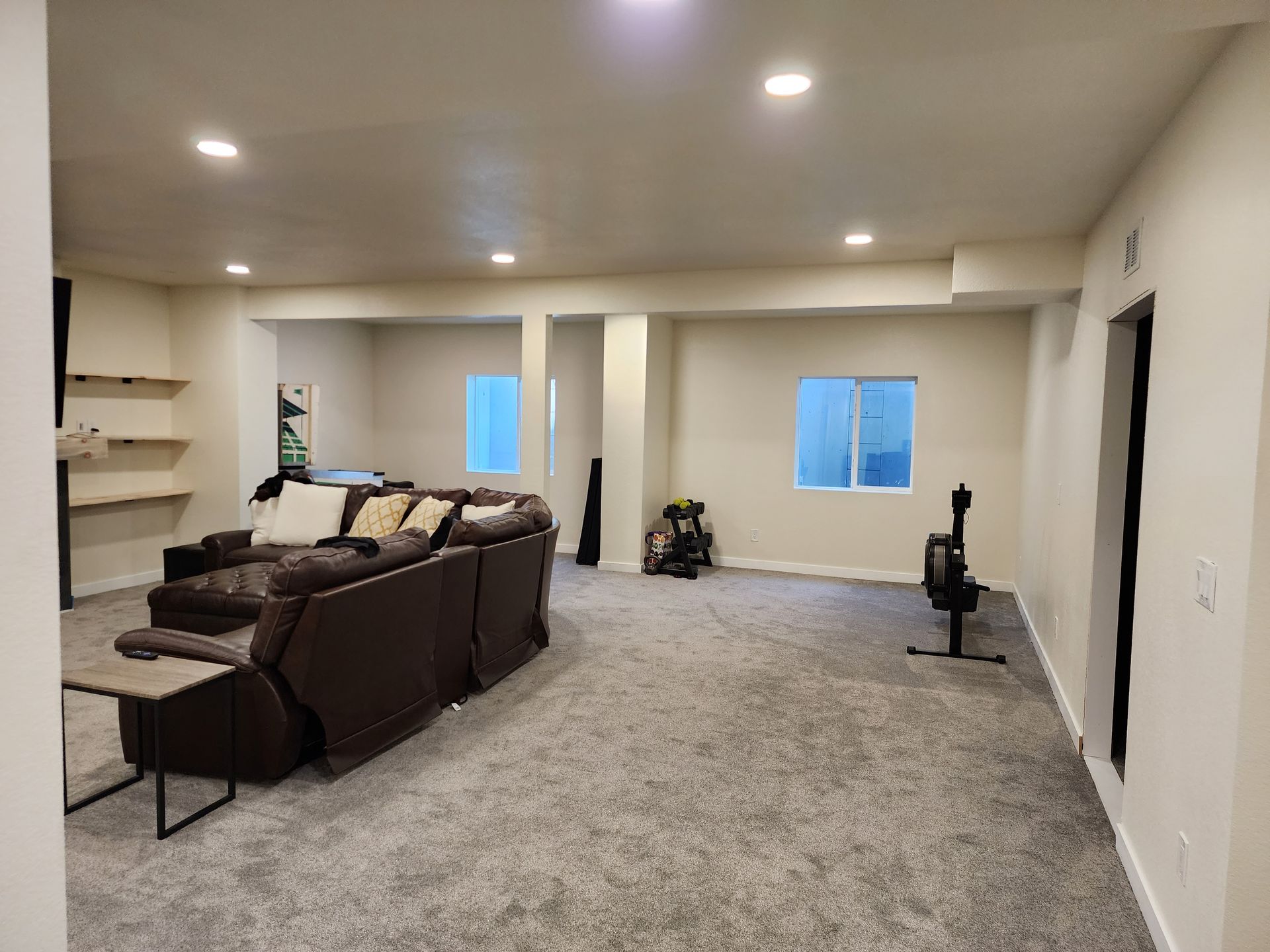Large finished basement with gray carpet, brown couch, and exercise equipment near windows.