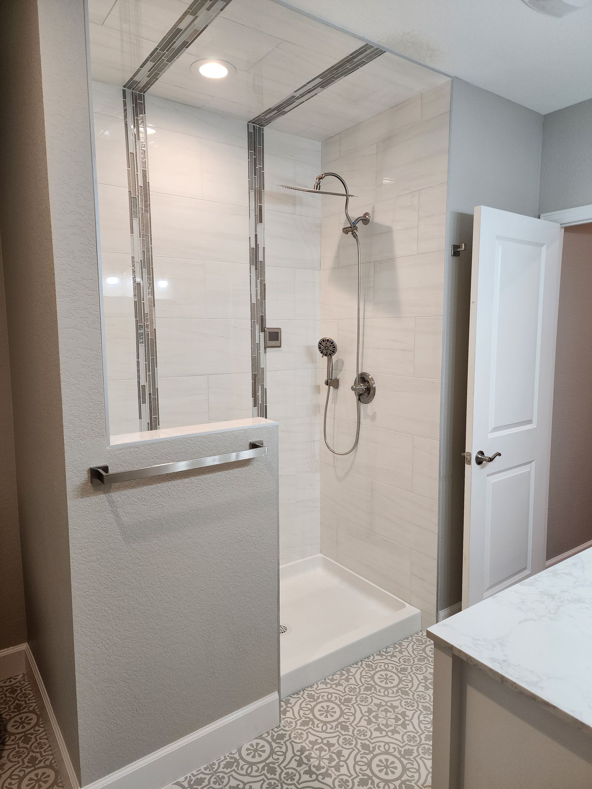 Modern, white tiled shower with glass door and silver fixtures. Grey walls, decorative floor tiles.