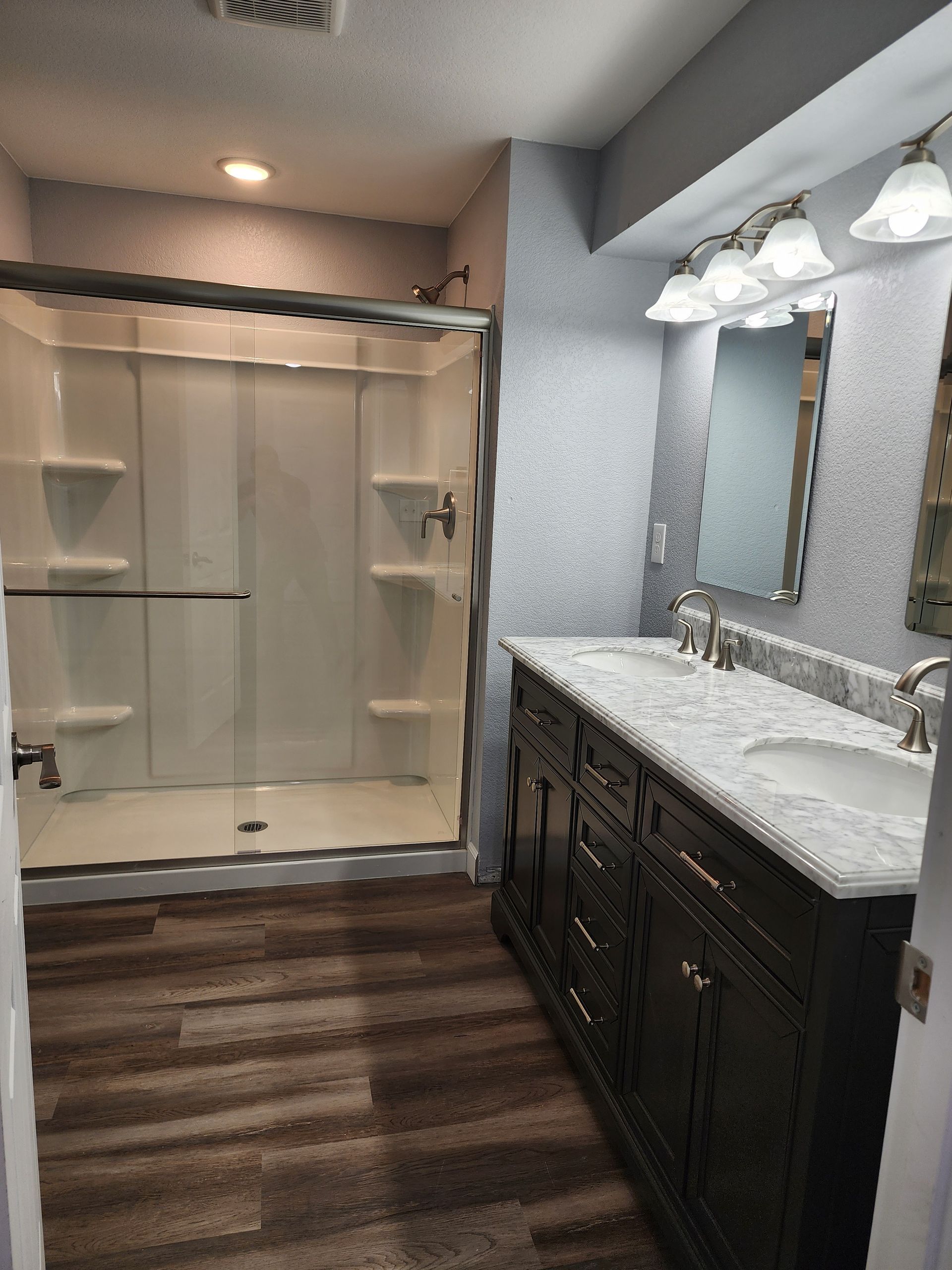 Bathroom with shower, double vanity, wood-look floor. Shower is white. Vanity is dark brown with white countertop.