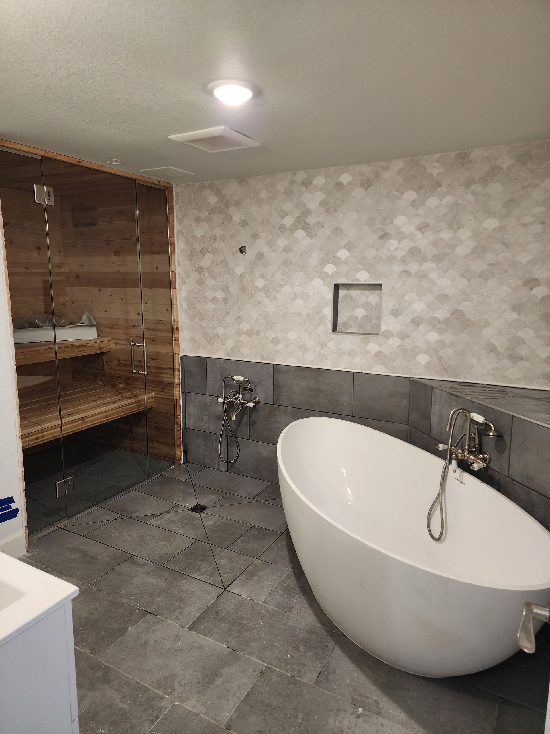Bathroom with white oval bathtub, gray tile floor and walls, wood sauna.