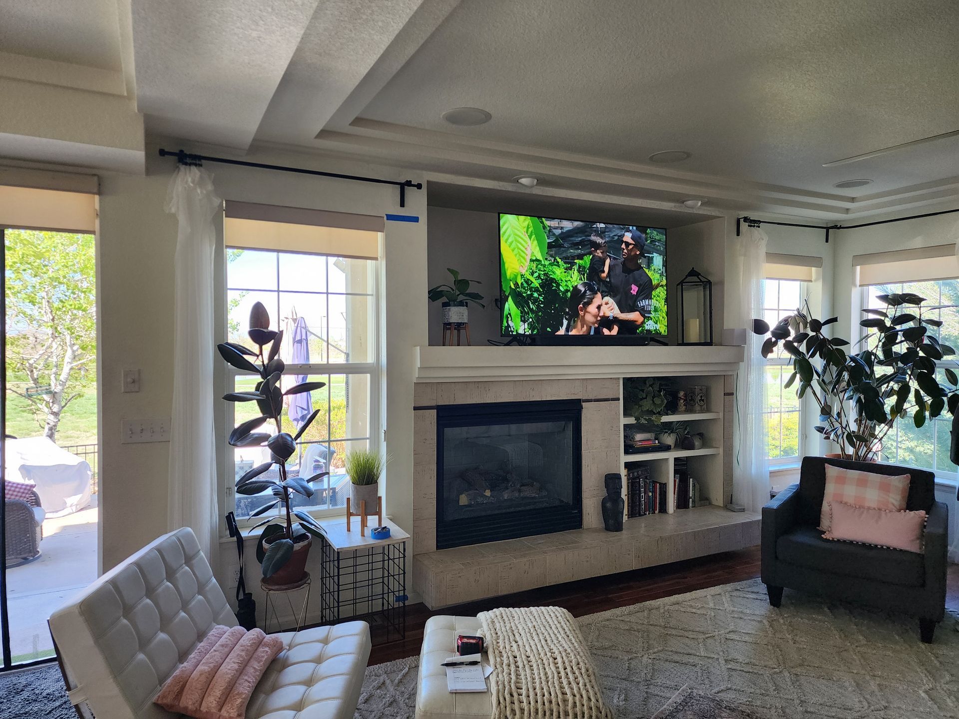 Living room with fireplace, TV, and seating; light-colored walls and curtains; sunlight streams in from the windows.
