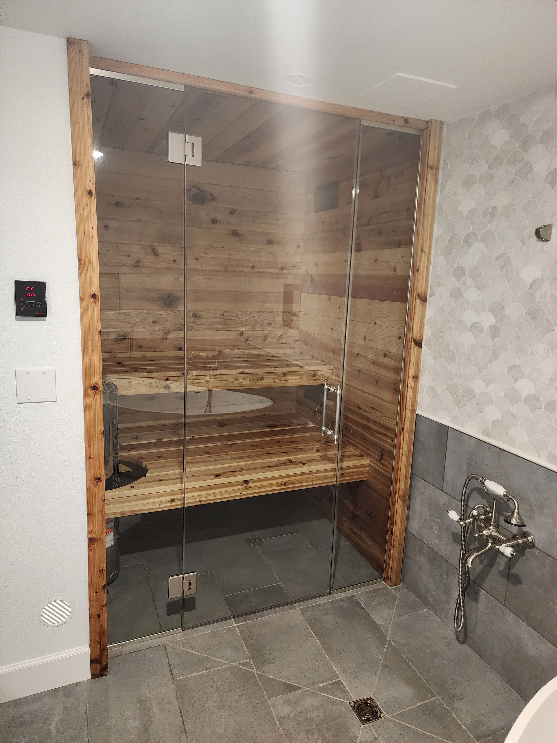 Glass-doored sauna with wood interior, built into a bathroom. Includes benches, a control panel, and a stone floor.
