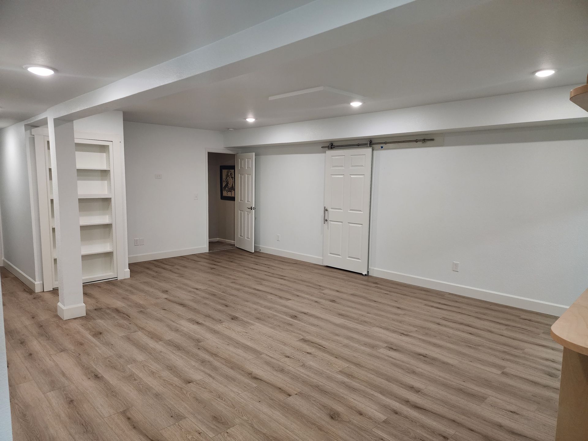 Empty basement room with light wood flooring, built-in shelves, and a sliding door.