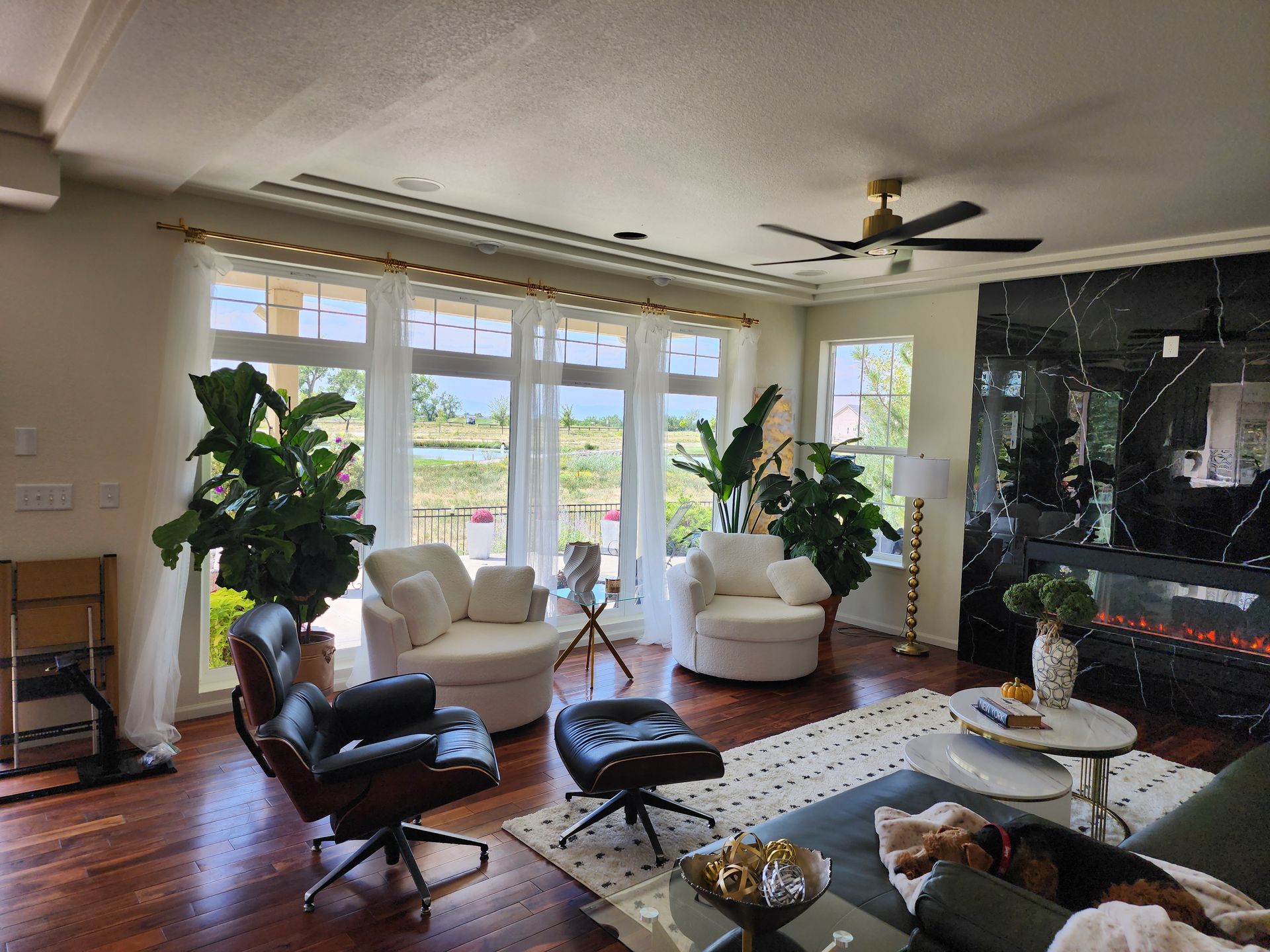 Living room with large windows, plants, and modern furniture. Features a black marble fireplace and white seating.