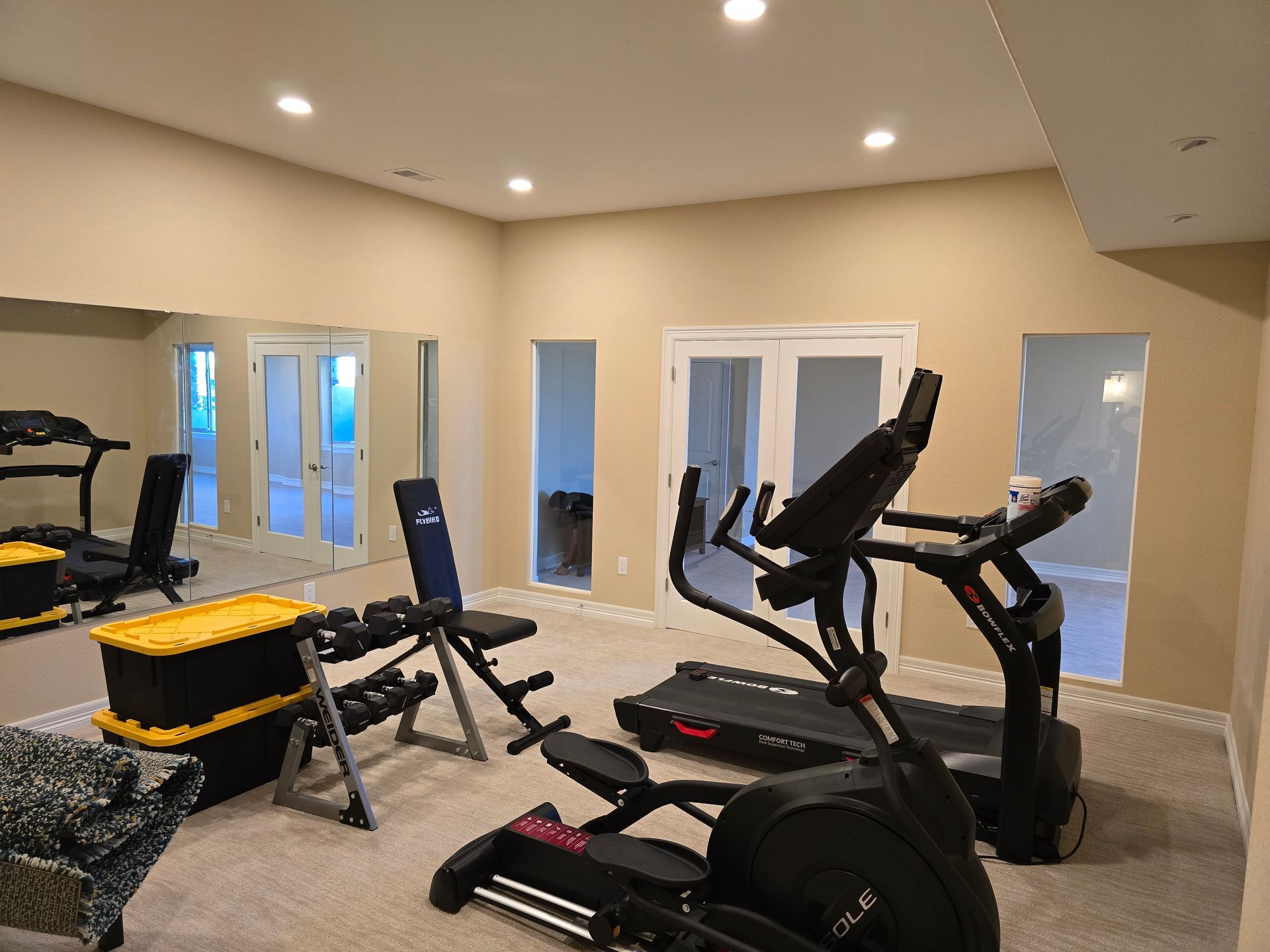 Home gym with exercise equipment: treadmill, elliptical, weights, and bench. Beige walls, mirror, and carpet.