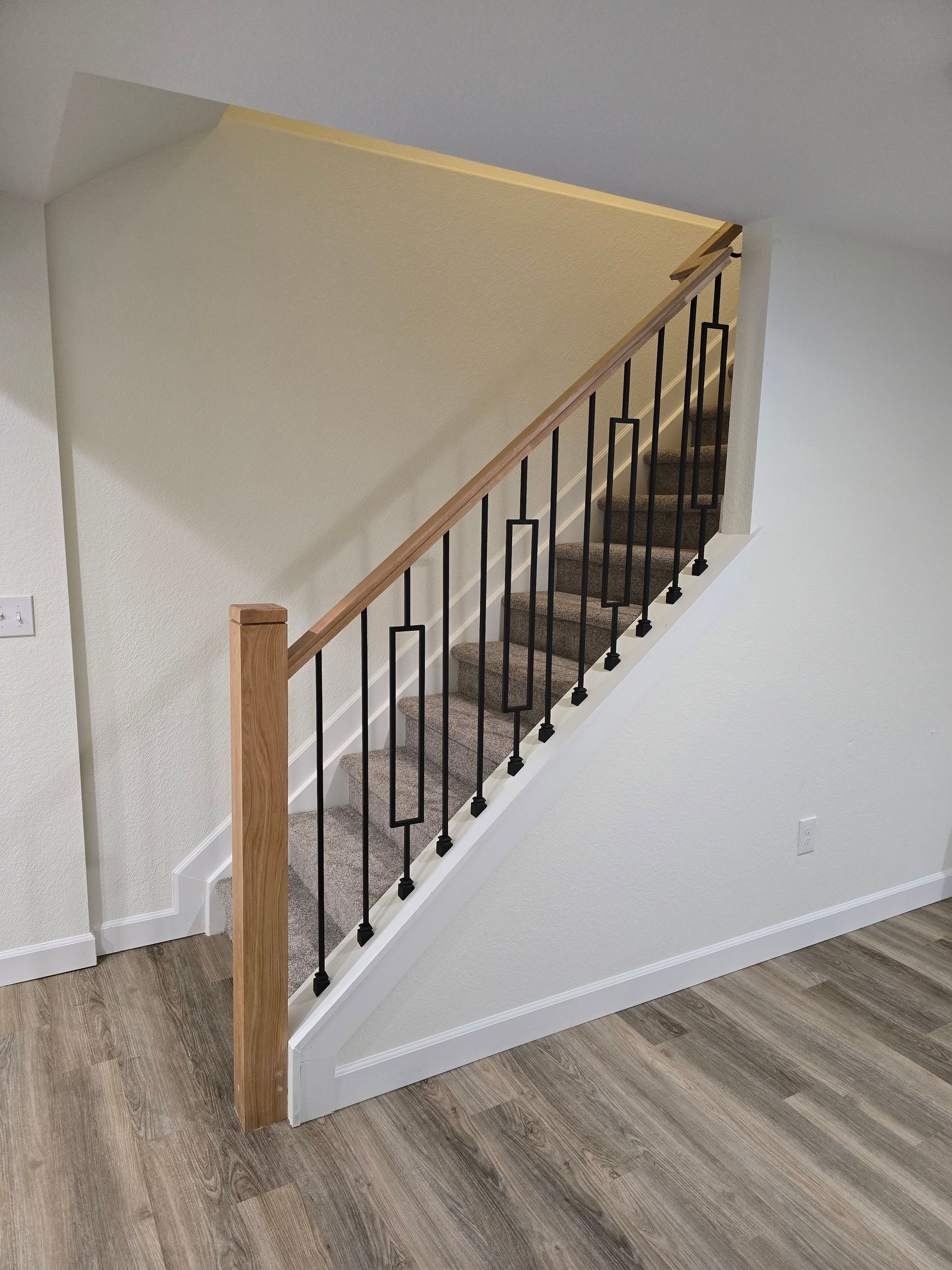 Staircase with wood handrail and black metal balusters. White walls, grey carpet, and wood-look flooring.