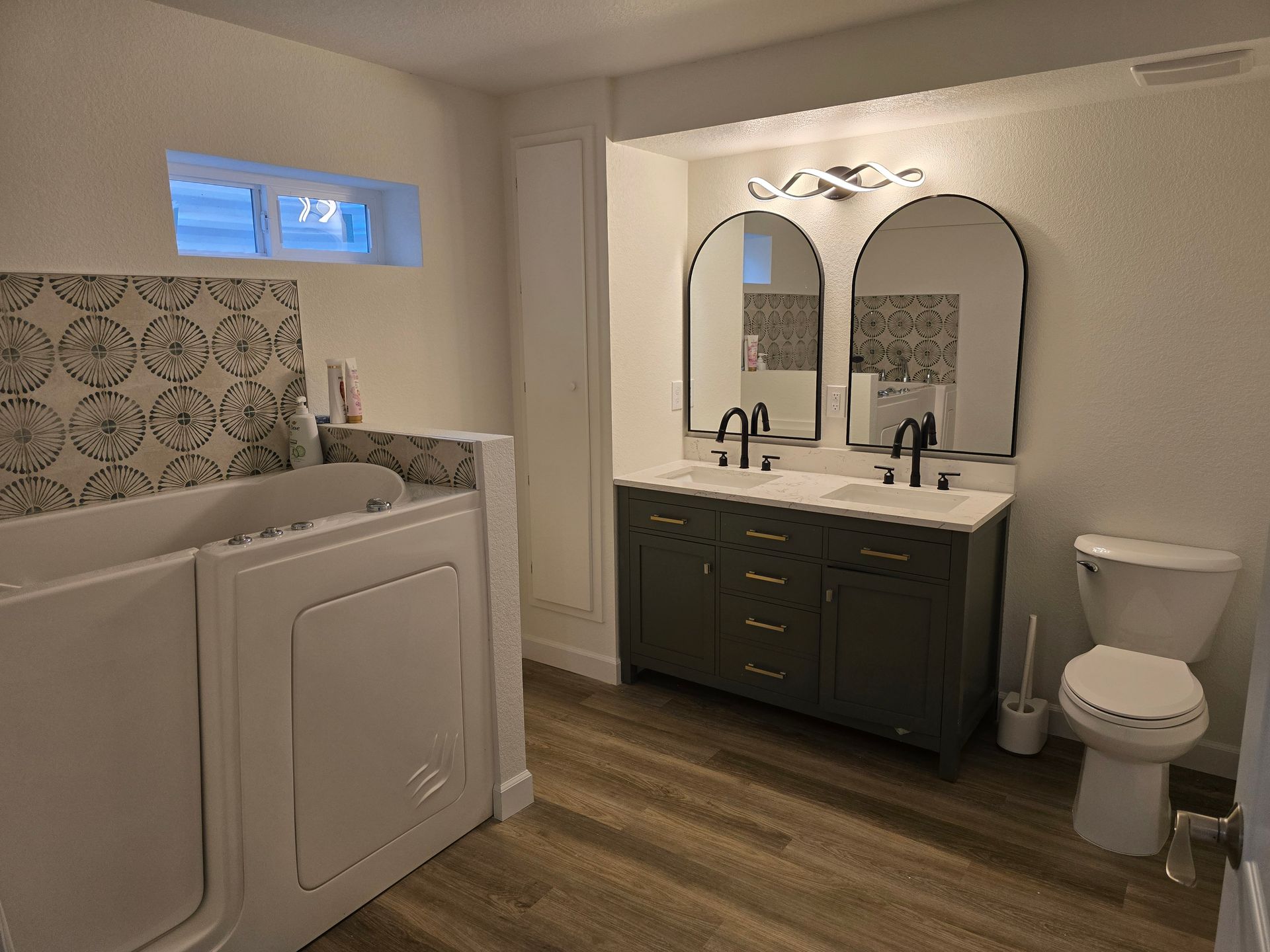 Bathroom with walk-in tub, dual sinks with arched mirrors, and a toilet. Neutral colors, wood-look flooring.
