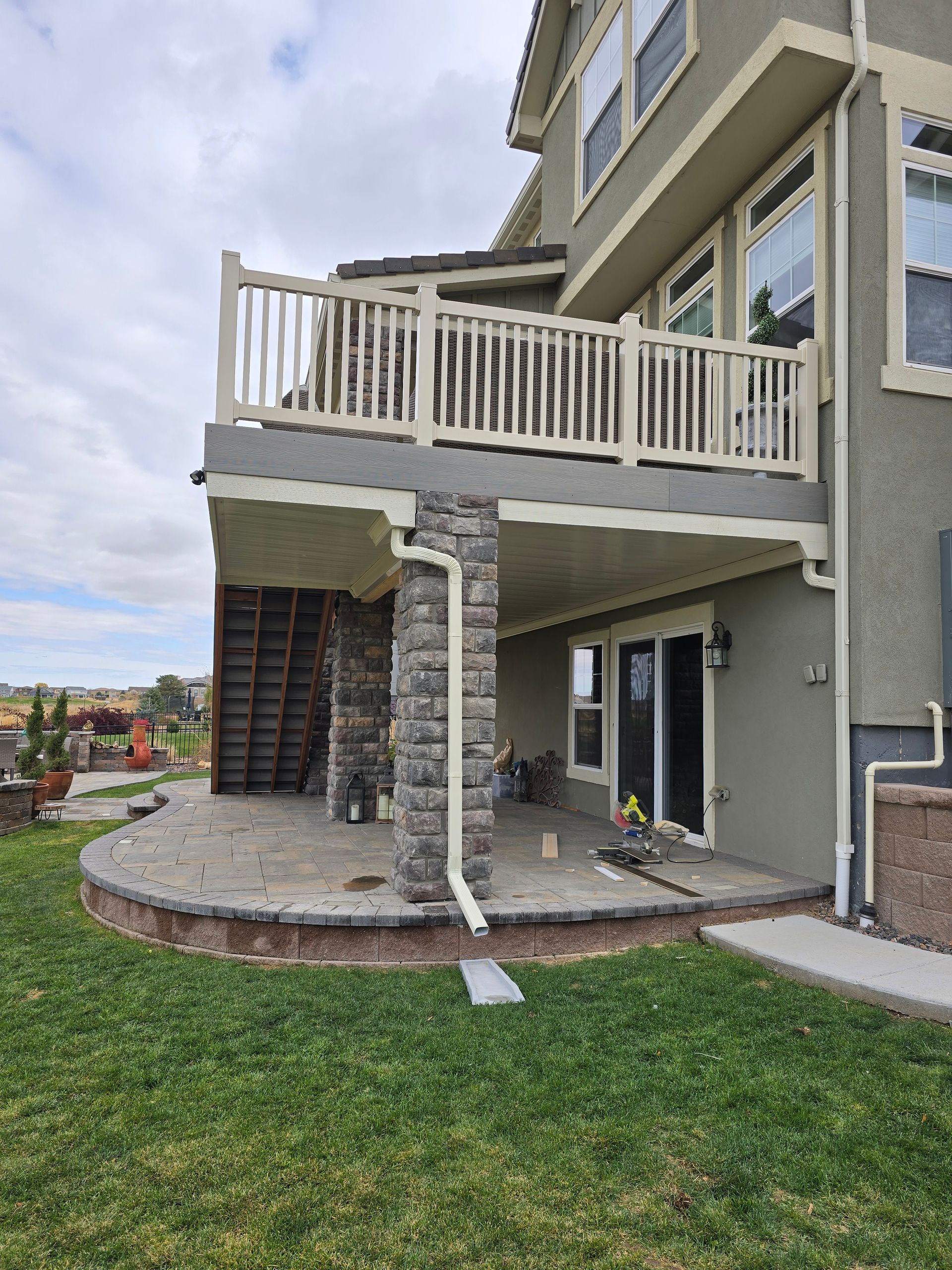 Two-story house with a deck. Stone-covered columns support the upper deck. Lower patio with grass and landscaping.