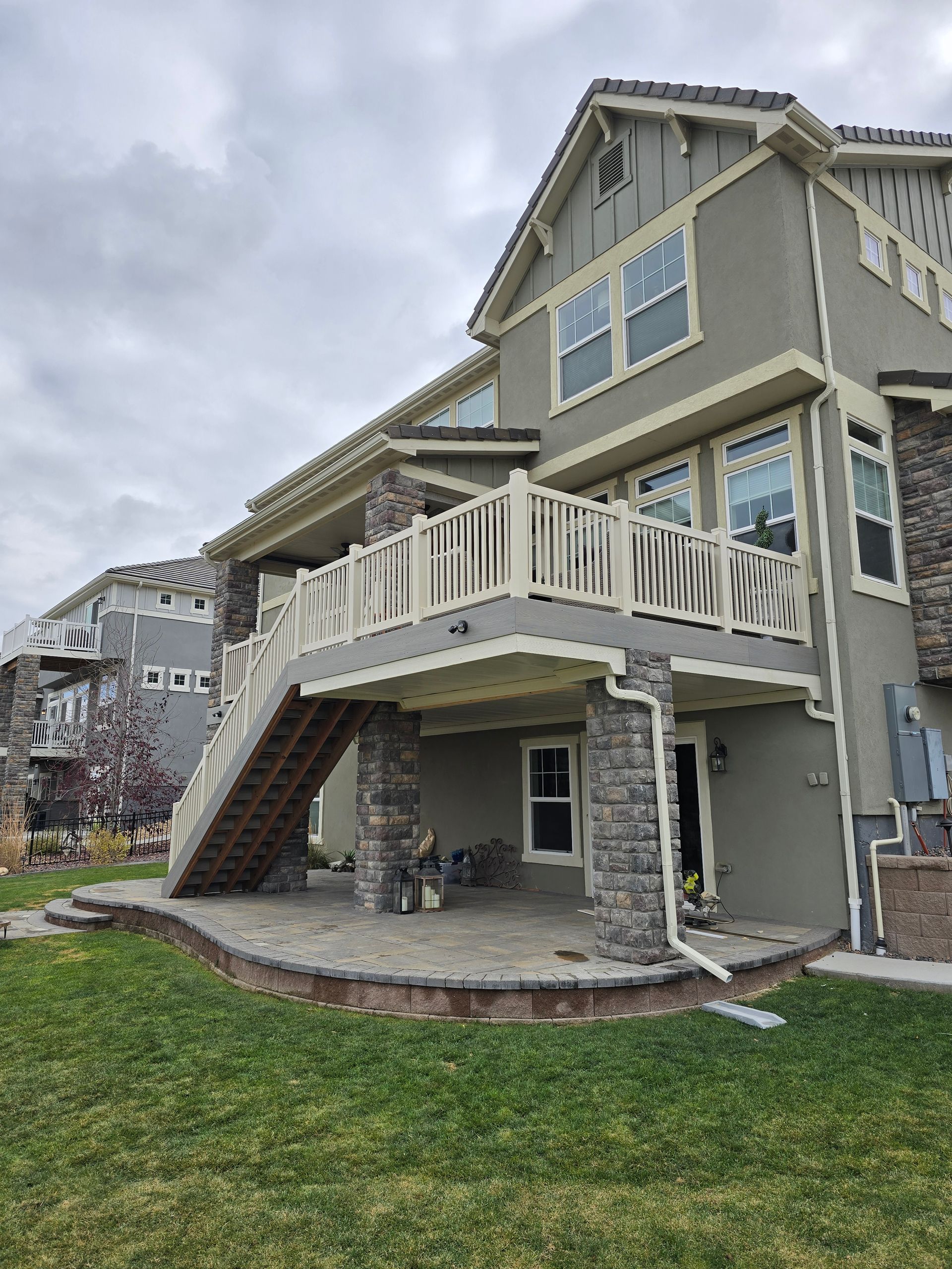 Two-story house with a deck, stairs, and stone columns, set on a grassy yard under an overcast sky.