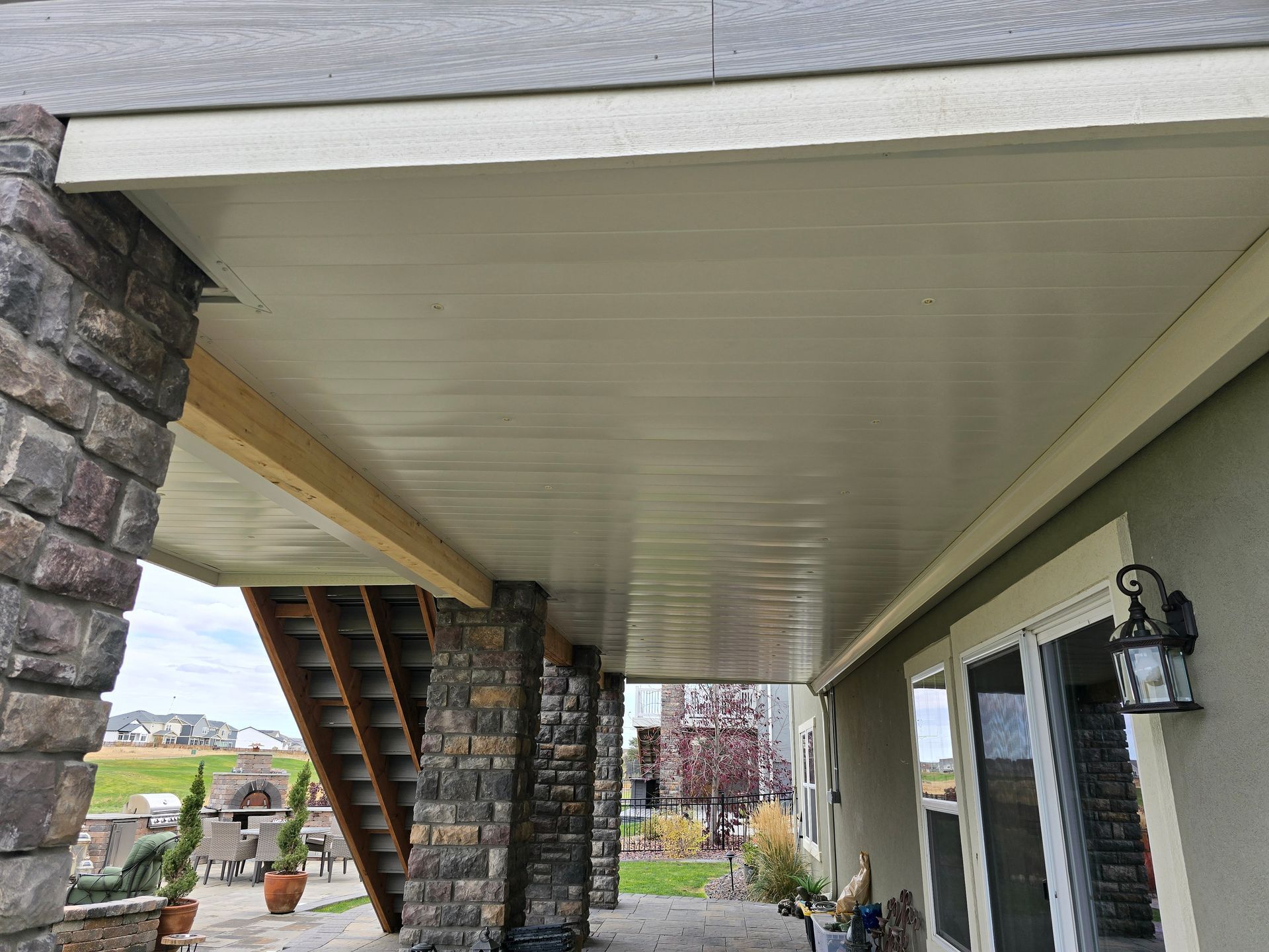 Beige soffit beneath a covered patio. Supports are stone and wood. House exterior visible.