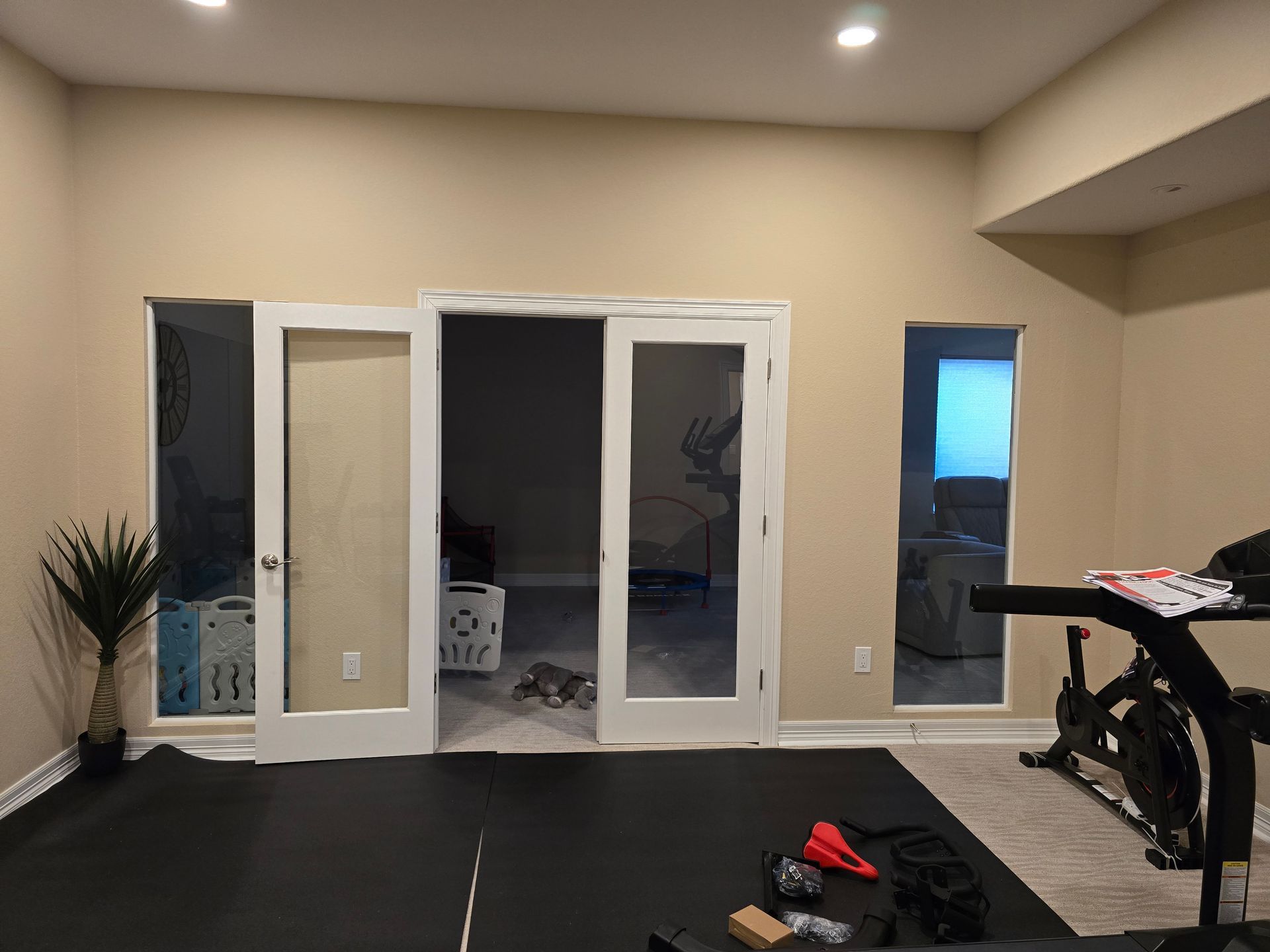 A home gym with a treadmill, French doors, and a large plant. Black mats cover the floor.