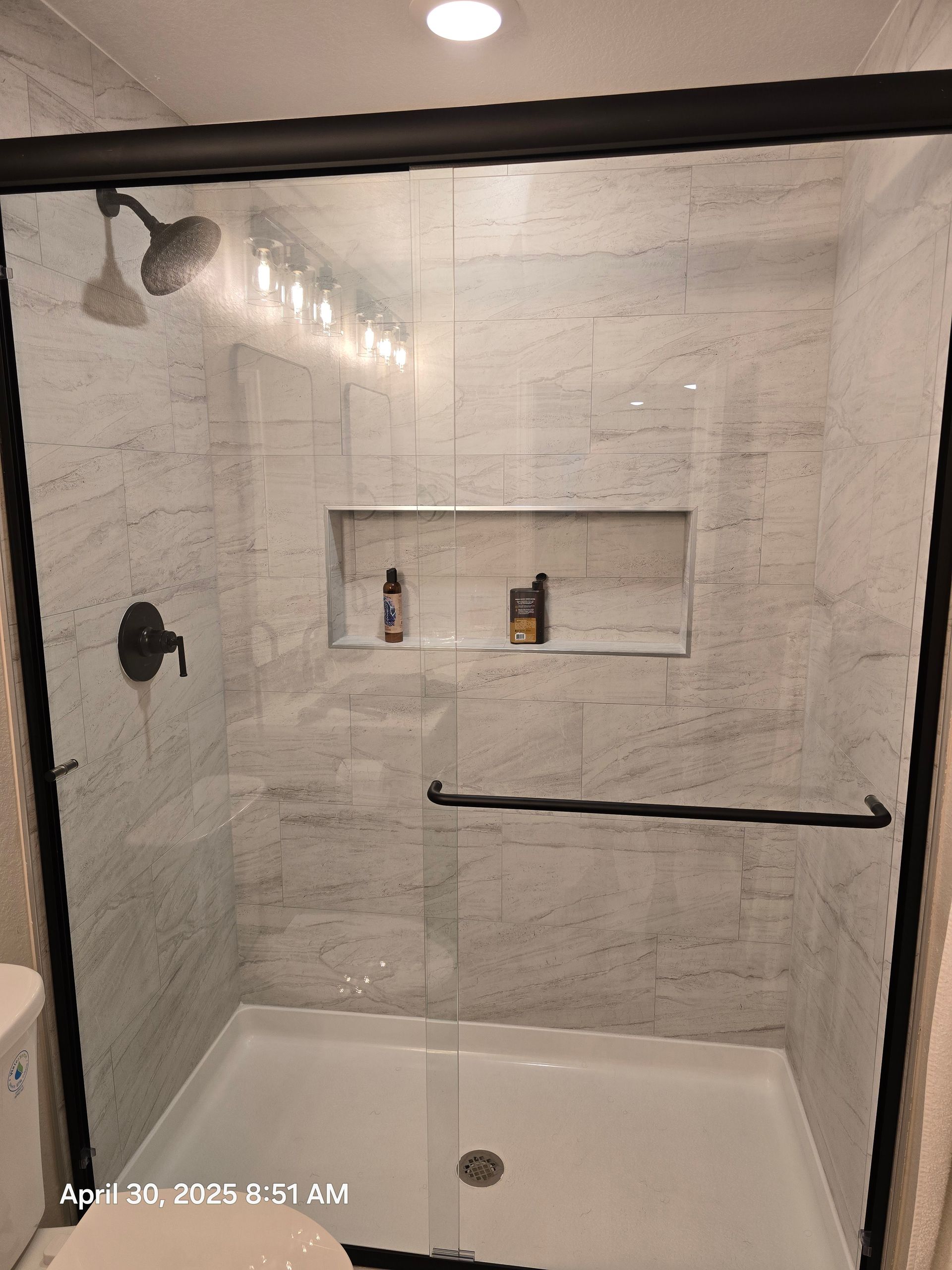 Bathroom with white tile walls, black shower door, fixtures, and built-in shelf with toiletries.